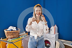 Young blonde woman at laundry room pointing fingers to camera with happy and funny face