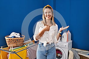 Young blonde woman at laundry room showing palm hand and doing ok gesture with thumbs up, smiling happy and cheerful