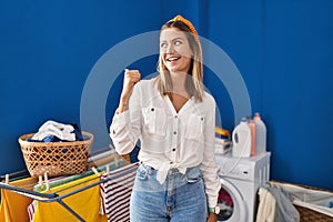 Young blonde woman at laundry room smiling with happy face looking and pointing to the side with thumb up