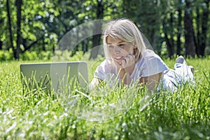 A young blonde woman lies in a park on the grass with a laptop. Blogging, education and remote work