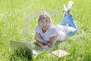 A young blonde woman lies in a park on the grass with a laptop. Blogging, education and remote work