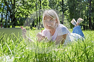 A young blonde woman lies in a park on the grass with a laptop. Blogging, education and remote work