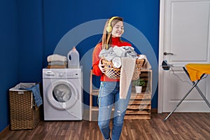 Young blonde woman listening to music holding basket with clothes at laundry room