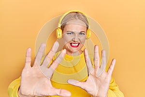 Young blonde woman listening to music using headphones afraid and terrified with fear expression stop gesture with hands, shouting