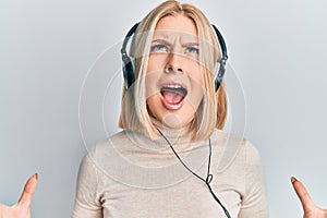 Young blonde woman listening to music using headphones crazy and mad shouting and yelling with aggressive expression and arms