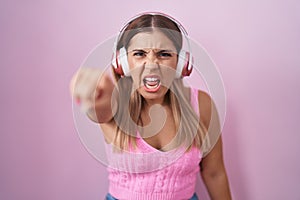 Young blonde woman listening to music using headphones pointing displeased and frustrated to the camera, angry and furious with