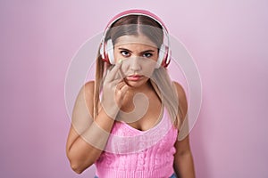 Young blonde woman listening to music using headphones pointing to the eye watching you gesture, suspicious expression