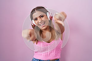 Young blonde woman listening to music using headphones pointing to you and the camera with fingers, smiling positive and cheerful