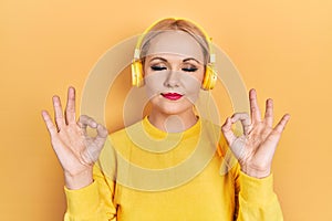 Young blonde woman listening to music using headphones relax and smiling with eyes closed doing meditation gesture with fingers