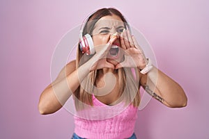 Young blonde woman listening to music using headphones shouting angry out loud with hands over mouth