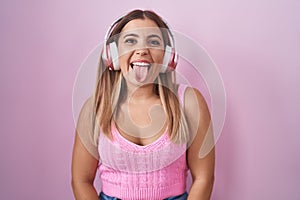 Young blonde woman listening to music using headphones sticking tongue out happy with funny expression