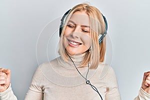 Young blonde woman listening to music using headphones very happy and excited doing winner gesture with arms raised, smiling and