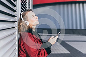 Young blonde woman listening to music in wireless headphones outdoors, holding mobile phone in her hands