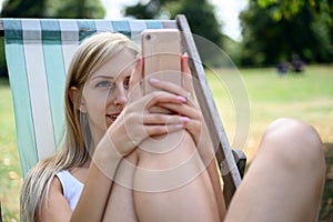 Young blonde woman in London Hyde Park, sitting in a beach chair and enjoying the late summer sun and browsing the internet on her