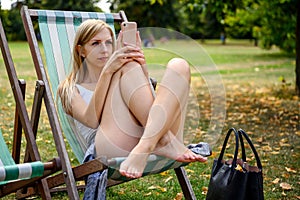 Young blonde woman in London Hyde Park, sitting in a beach chair and enjoying the late summer sun and browsing the internet on her