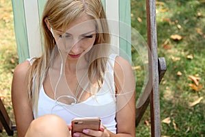 Young blonde woman in London Hyde Park, sitting in a beach chair and listening to music online on her mobile phone