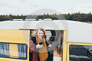 Young blonde woman looking out of camper van with solar panel on the roof top and pine forest on the background