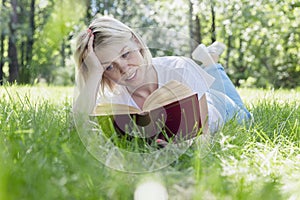 A young blonde woman is lying on the grass in a park on a sunny summer day and is reading a book. Recreation and distance learning