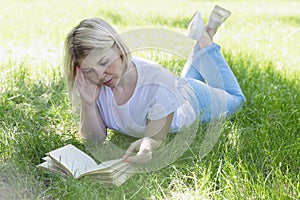 A young blonde woman is lying on the grass in a park on a sunny summer day and is reading a book. Recreation and distance learning