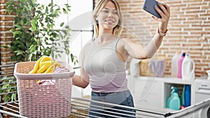 Young blonde woman make selfie by smartphone hanging clothes on clothesline at laundry room