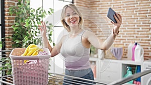 Young blonde woman make selfie by smartphone hanging clothes on clothesline at laundry room