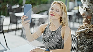 Young blonde woman making selfie by the smartphone sitting on table at coffee shop terrace