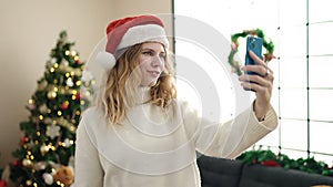 Young blonde woman making selfie by smartphone standing by christmas tree at home