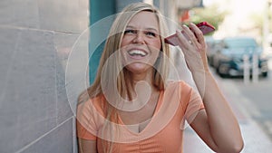 Young blonde woman miling confident listening audio message by the smartphone at street