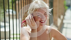 Young blonde woman miling confident listening audio message by the smartphone at street