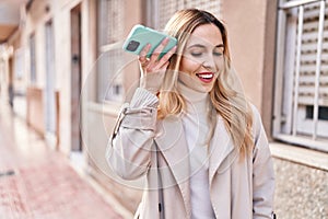 Young blonde woman miling confident listening audio message by the smartphone at street