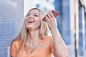 Young blonde woman miling confident listening audio message by the smartphone at street