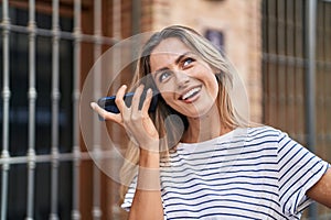 Young blonde woman miling confident listening audio message by the smartphone at street