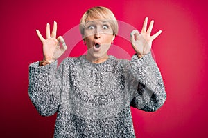 Young blonde woman with modern short hair wearing casual sweater over pink background looking surprised and shocked doing ok