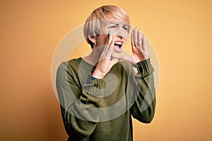 Young blonde woman with modern short hair wearing casual sweater over yellow background Shouting angry out loud with hands over