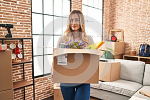 Young blonde woman moving to a new home holding cardboard box clueless and confused expression