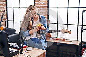 Young blonde woman musician playing electrical guitar using smartphone at music studio
