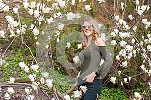 Young blonde woman near blossoming magnolia flowers tree in spring park on sunny day. Magnolia trees. Beautiful happy