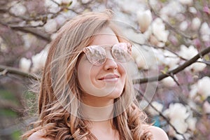 Young blonde woman near blossoming magnolia flowers tree in spring park on sunny day. Magnolia trees. Beautiful happy girl