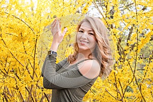 Young blonde woman near blossoming yellow trees in spring park on sunny day. Beautiful happy girl enjoying smell in a