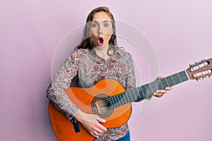 Young blonde woman playing classical guitar afraid and shocked with surprise and amazed expression, fear and excited face