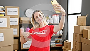 Young blonde woman pointing to volunteer uniform making selfie by smartphone at charity center