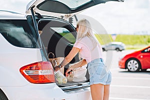 Young blonde woman put her shopping eco bags with food into car trunk on a parking.