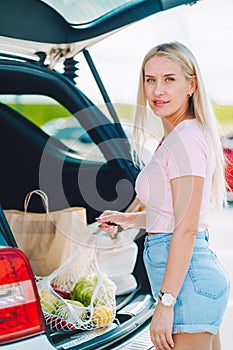 Young blonde woman put her shopping eco bags with food into car trunk on a parking.