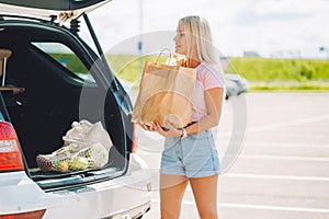 Young blonde woman put her shopping eco paper bags into car trunk on a parking.