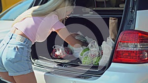 Young blonde woman put her shopping eco paper bags into car trunk on a parking