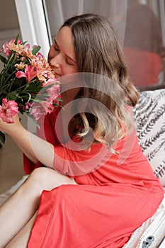 Young Blonde woman in a red robe with flowers against wide window background having lazy morning