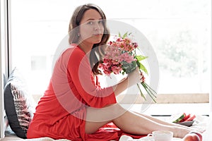Young Blonde woman in a red robe with flowers against wide window background having lazy morning