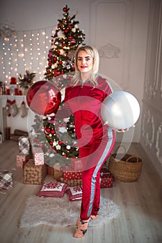Young blonde woman in a red tracksuit on Christmas decorations, on the background of a Christmas tree. Big balls, lights, garlands
