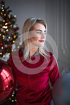 Young blonde woman in a red tracksuit on Christmas decorations, on the background of a Christmas tree. Big balls, lights, garlands