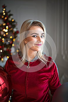 Young blonde woman in a red tracksuit on Christmas decorations, on the background of a Christmas tree. Big balls, lights, garlands